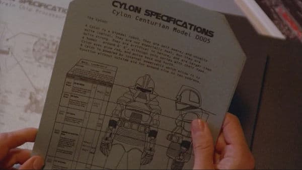 Screengrab from Battlestar Galactica showing the diagram of a cylon, the main antagonist robot from the series. The diagram is on a piece of 8-inch by 11-inch paper with 45-degree angular corners on the page. There's a black manilla folder behind the piece of paper that also has its corners cut to the same angle.