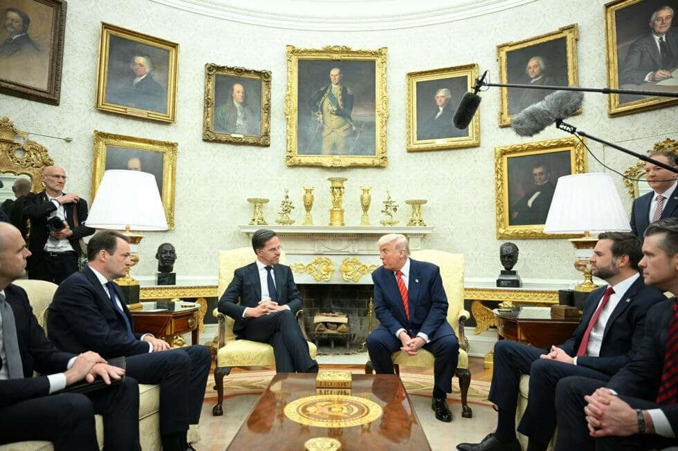 Donald Trump in the Oval Office meeting with a head of state. The two presidents sit in the center of the frame with the Vice President and other cabinet members forming a v-shape extening towards the camera. At the top of the frame boom microphones dangle. Behind Trump is the fireplace, now adorned in gold finishes, with gold urns on top of the mantle, and gold borers around all the picture frames (except Teddy Roosevelt). The side tables with bronze busts are also encrusted with gold. The lamps gold. The mirror gold.